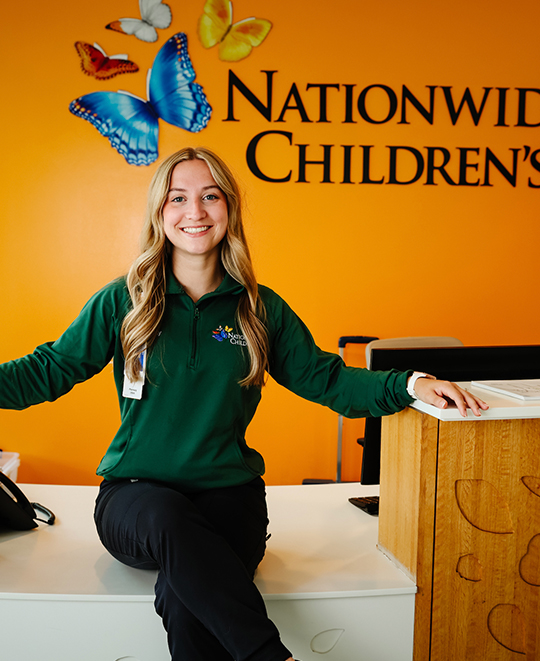 Photo of Ashley sitting on a desk in front of sign on a wall saying Nationwide Childrens 