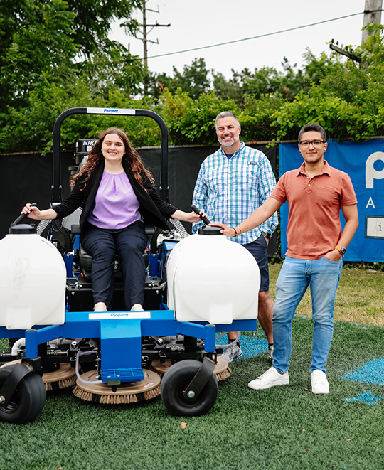 Photo of Ella sitting on a machine that paints fields posing with 2 other people