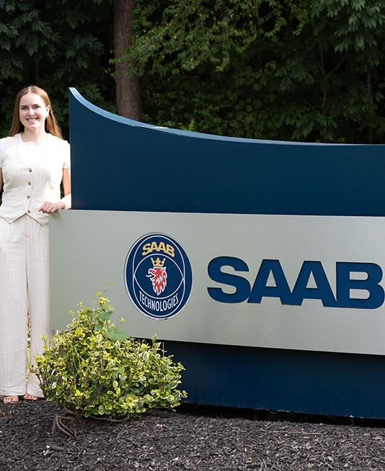 Photo of Rylie standing outside next to a SAAB sign 