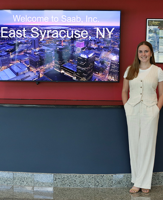 Photo of Rylie smiling next to 'Welcome to SAAB, Inc.' sign