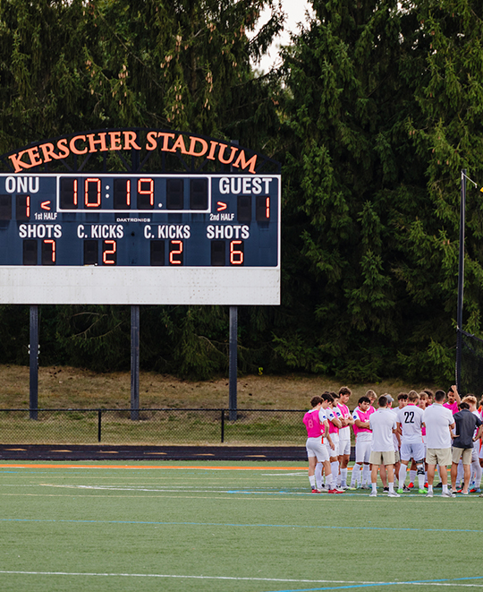 Photo of ONU men soccer team on the sidelines discussing, with the scoreboard in the background with a tie of 1 to 1