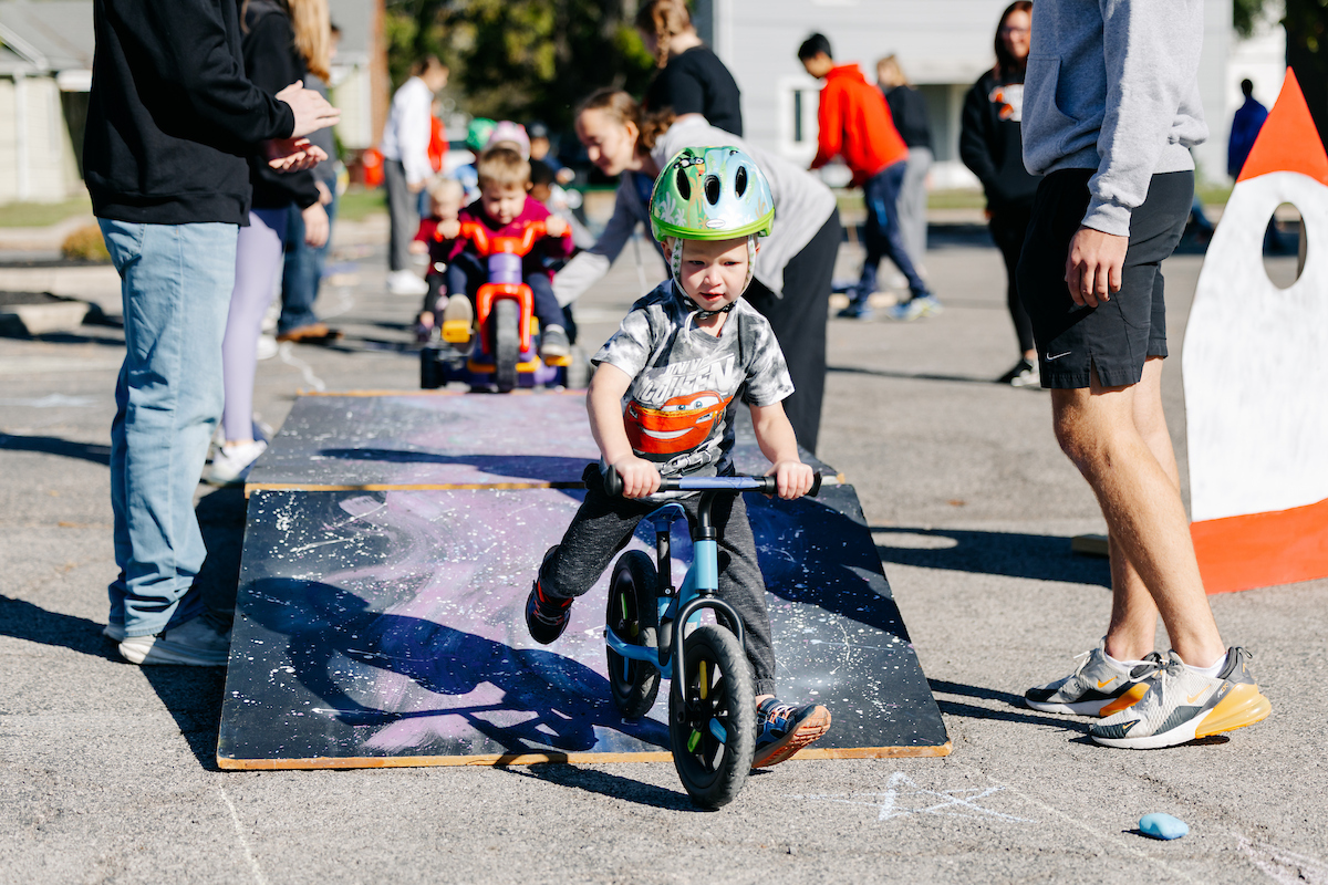 A small child riding a bicycle down a wooden ramp in a parking lot