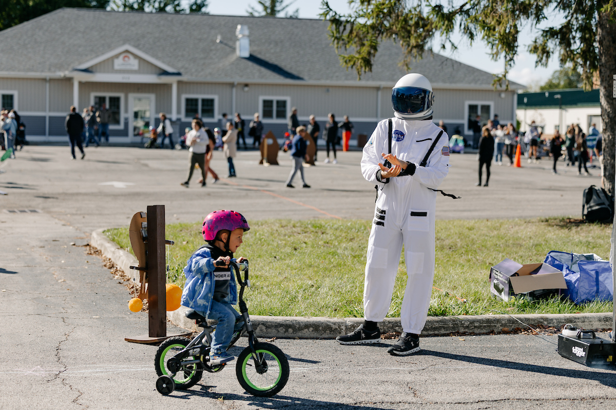 A small child riding a bicycle past a person dressed as an astronaut in a parking lot