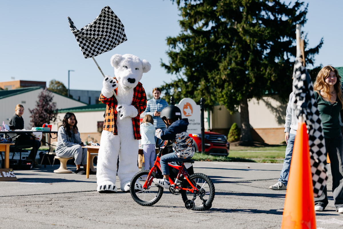 A polar bear mascot waves a checkered flag as a child rides a bicycle