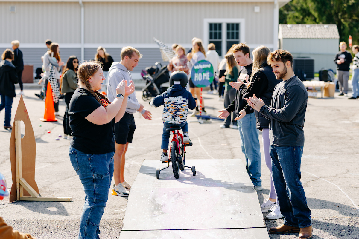 A small child riding a bicycle over a wooden ramp in a parking lot