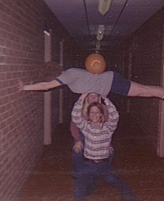 Old Photo of hallway shenanigans with a couple student holding a perfectly horizonal friend above their head with a pumpkin resting on their back.