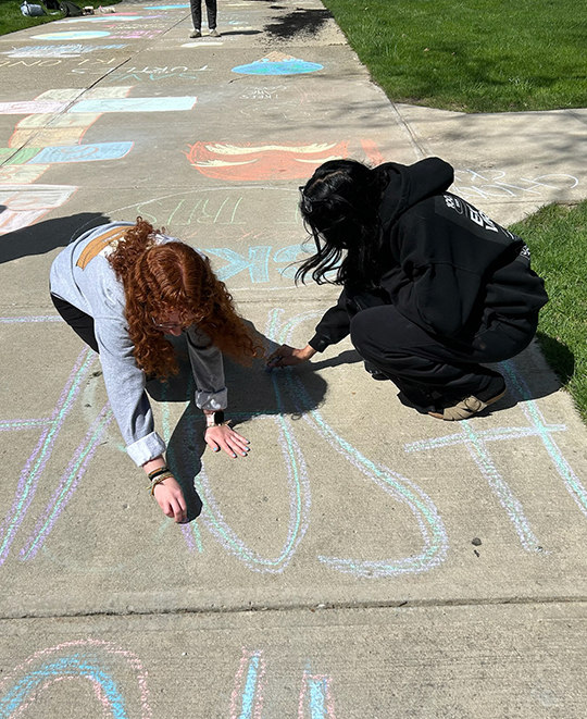 2 students on hands and knees on the sidewalk using chalk to write