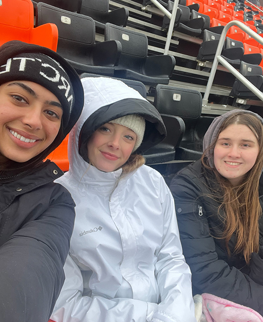 Selfie of 3 gals dressed warmly in jackets seated in bright orange football stadium seats