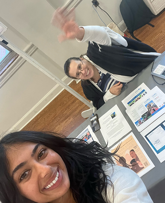 Selfie where there is a woman in the background seated behind a table full of papers waving