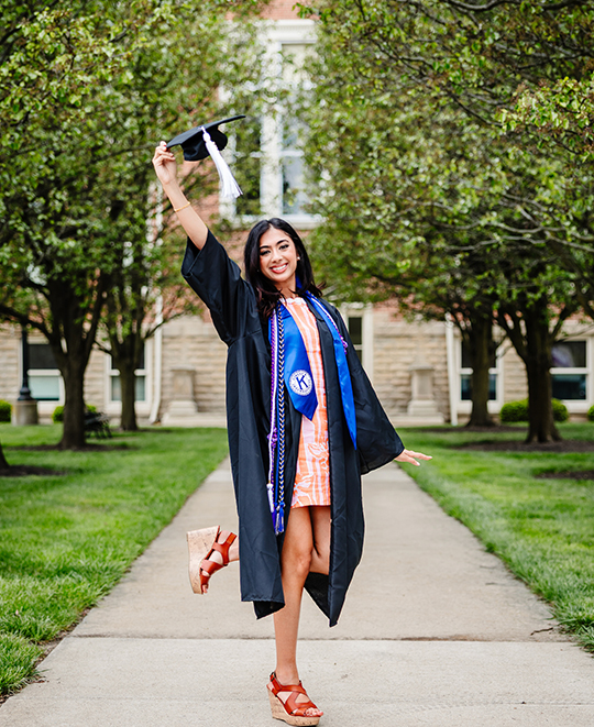 Simran in her graduation gown raising cap in the air posing on a sidewalk with grass and trees beside her.