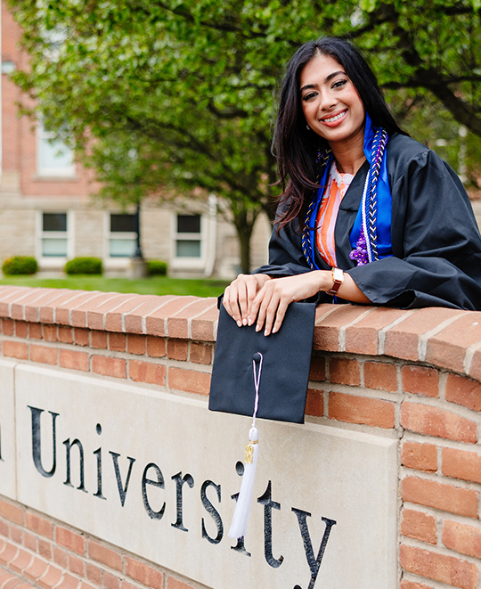 Simran in her graduation cap and gown posing behind the ONU brick outdoor sign