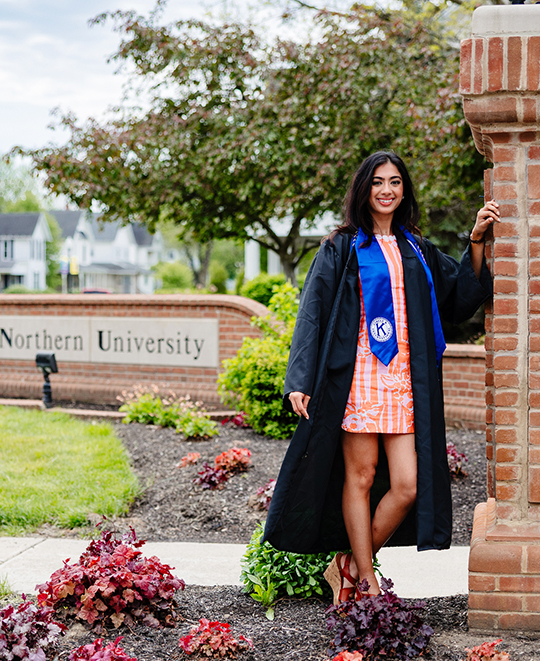 Simran in her graduation cap and gown standing beside tall brick pillar in front of ONU brick outdoor sign