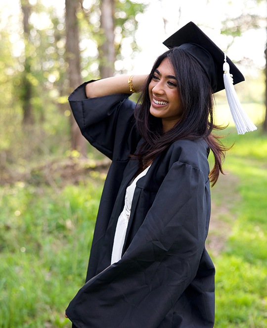 Simran in her graduation cap and gown smiling in a grassy field