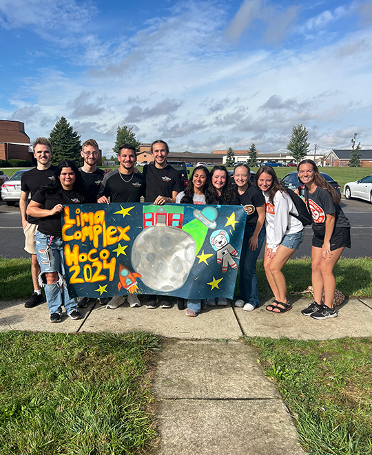 Big group on a sidewalk holding a banner that says Lima complex hoco 2024 and a moon and anstronaut