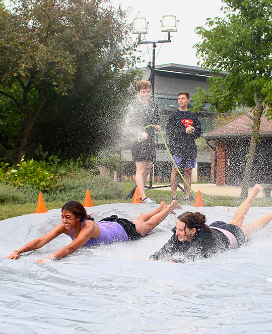 2 females on a slip on slide with background students hosing it down