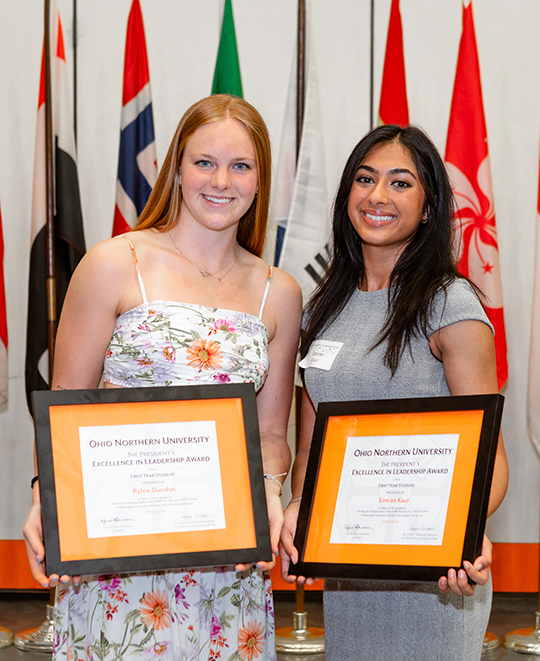 2 females holding awards -Excellence leadership award - posing together with a collection of flags behind them