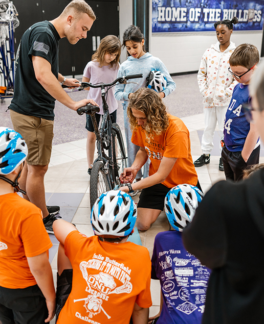 Adult male in the middle of a circle of children seated watching as he demonstrates how to work with the bike