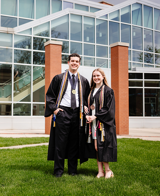 Drew and Rylie posing in their graduation gowns posing in front of Dicke Hall 