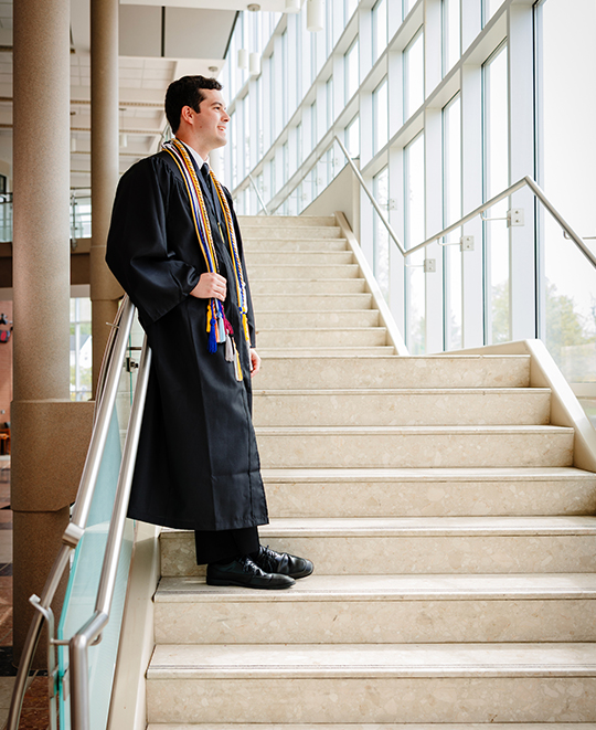 Drew in his graduation gown, standing in a stair case looking out the windows
