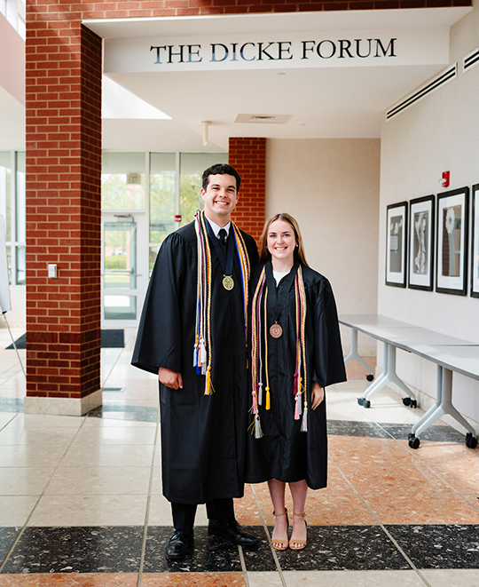 Drew and Rylie posing in their graduation gowns posing underneath a hallway sign that says the Dicke Forum