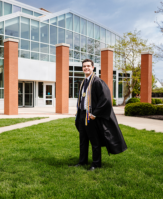 Drew posing in his graduation gowns posing in front of Dicke Hall 