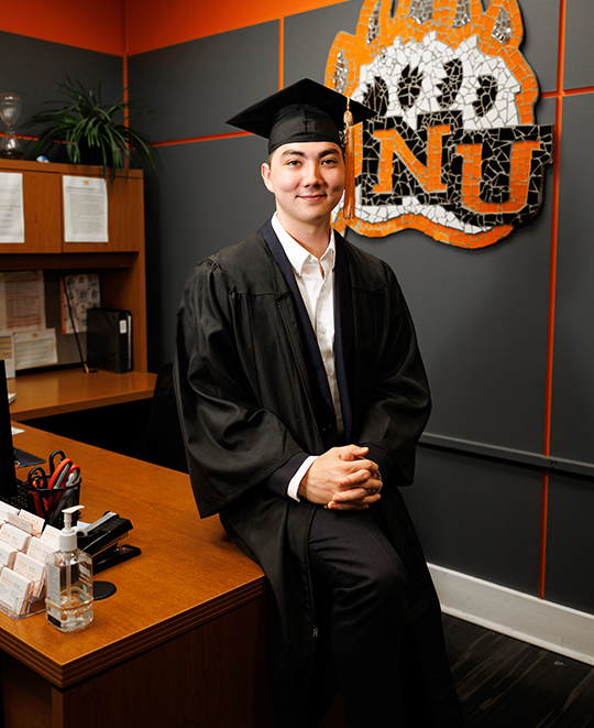 Dylan leaning on a desk in a gray room with ONU bear paw logo on the wall