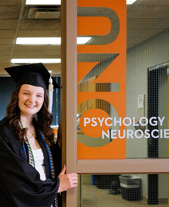 Julia standing in the ONU psychology doorway in her graduation cap and gown