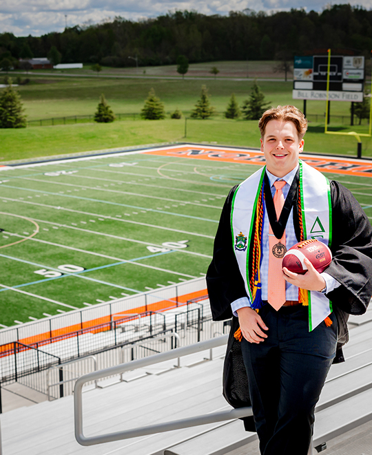 Jordan in his graduation gown holding a football at the top of the stadium with the football field in the background