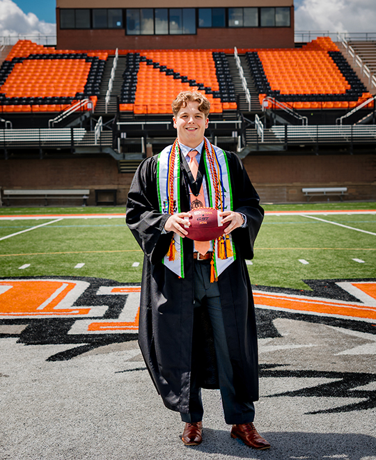 Jordan in his graduation gown holding a football posing at the center field