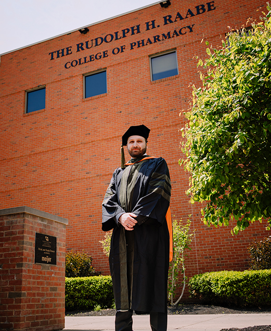 Joy wearing his graduation gown posing outside the college of pharmacy building