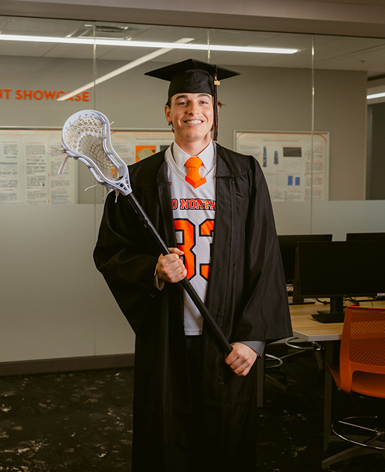 Mark wearing his graduation cap and gown holding his lacrosse stick