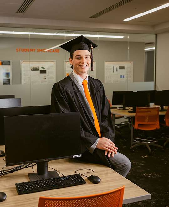 Mark seated on a desk next to a computer wearing a cap and gown