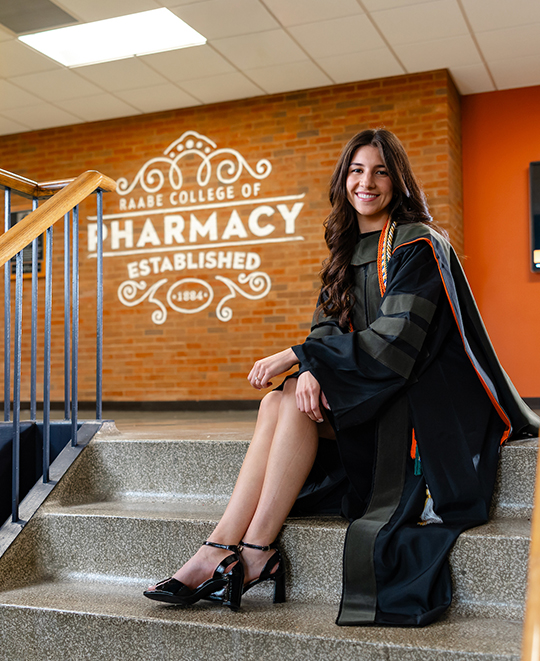 Samantha seated on the top steps of a staircase with a pharmacy sign on the wall in the background