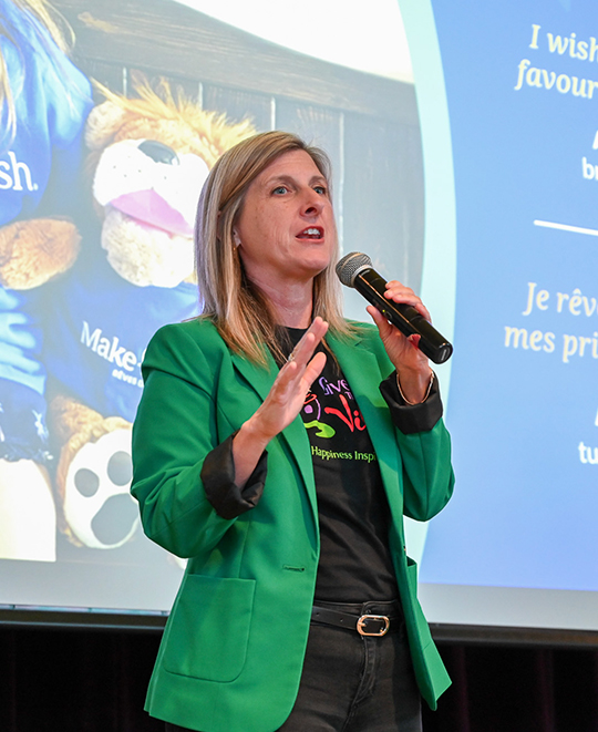 Caroline Schumacher, wearing a vibrant green blazer and holding a microphone, speaks passionately on a stage with a colorful, animated presentation displayed on the screen behind her.