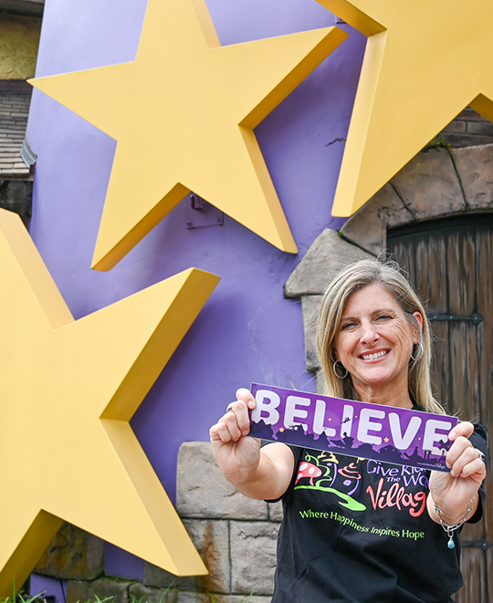 A close-up portrait of Caroline Schumacher smiling next to large, yellow decorative stars, holding up a small sign that reads "BELIEVE."