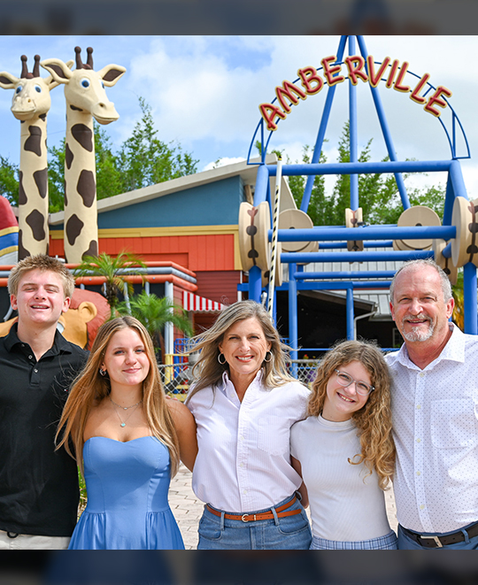 Caroline Schumacher smiles alongside a group of adults and children as they stand together in front of the whimsical, colorful entrance of Give Kids The World Village.