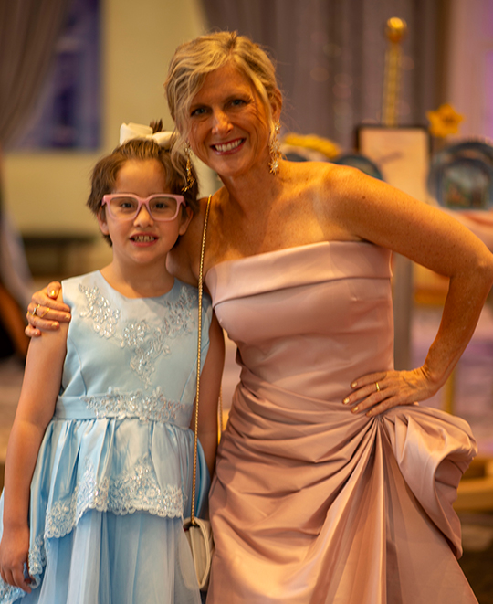 Caroline Schumacher, wearing an elegant strapless gown, stands smiling with her arm around a young girl in a blue dress during a formal evening event.