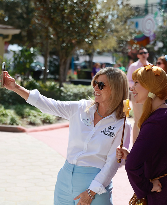 Caroline Schumacher, wearing a white button-down shirt and light blue pants, stands outside smiling and gesturing with her hand while talking with another woman.