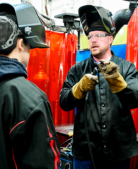 A manufacturing technology teacher in safety glasses and a black shirt converses with a student wearing a welding helmet in a high school workshop setting, with red welding privacy curtains visible in the background.