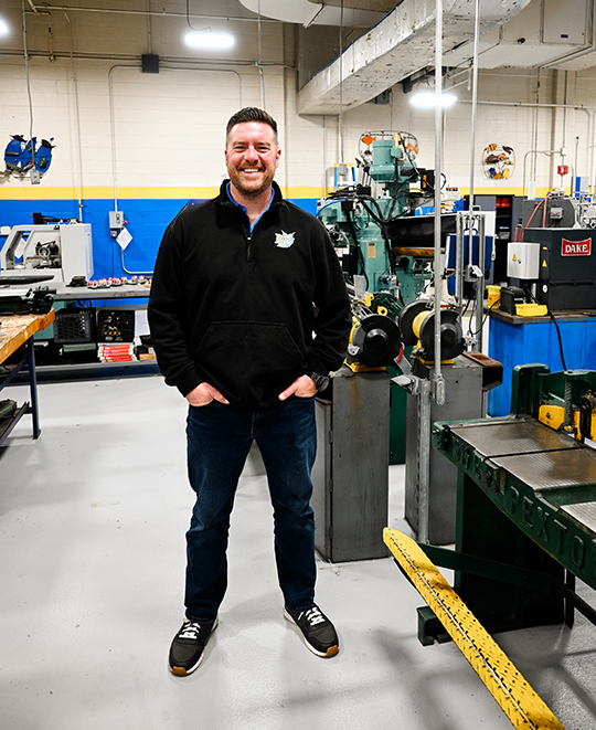 A full-length portrait of manufacturing technology teacher David Mason standing in a high school workshop, smiling while wearing safety glasses, a black pullover shirt, and jeans, with various machining equipment visible in the background.