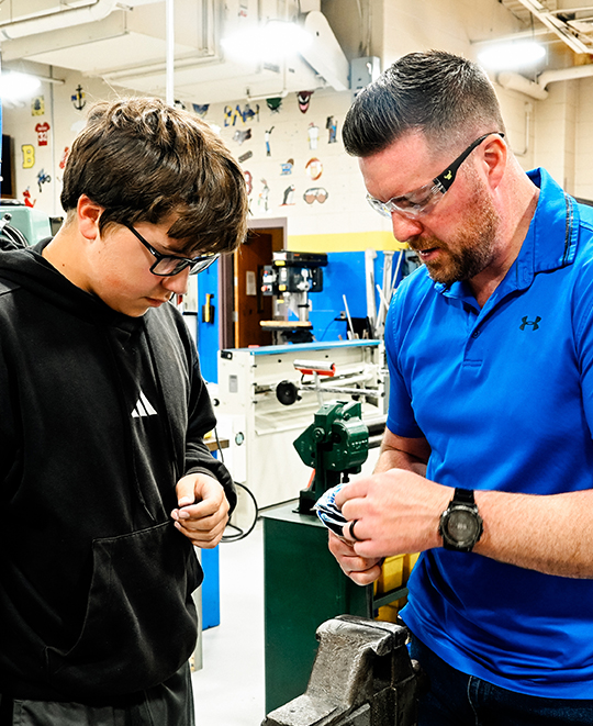 Manufacturing technology teacher David Mason, wearing a blue polo shirt and safety glasses, works closely with a student in a high school workshop, guiding them through a hands-on project with machinery visible in the background.