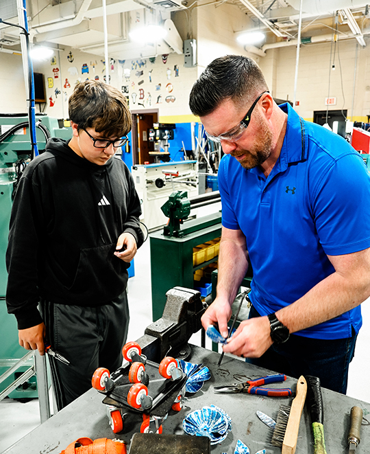 Manufacturing technology teacher David Mason, wearing a blue polo shirt and safety glasses, stands beside a student in a high school workshop as they look down and collaborate on a hands-on technical project.