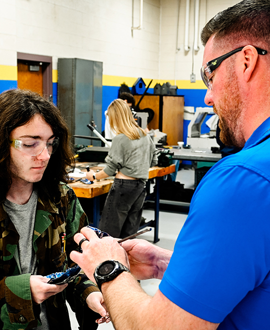 Manufacturing technology teacher David Mason, wearing safety glasses and a blue polo shirt, stands in a workshop alongside a student as they examine a small component together.
