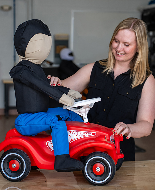 Taylor Weideling smiling alongside a crash test dummy mannequin and a red toy car.