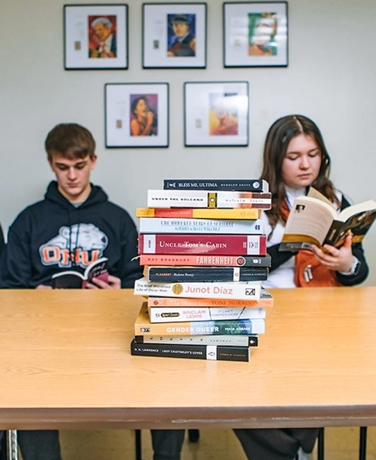 A stack of books on a table with 2 students in the background reading