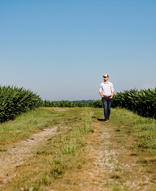 Alumnus Dr. Eric Schultz standing along a grassy path next to an agricultural research crop field under a clear blue sky.