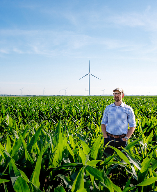Alumnus Dr. Eric Schultz standing in an agricultural research field, showcasing tall crops and wind turbines under a bright sky.