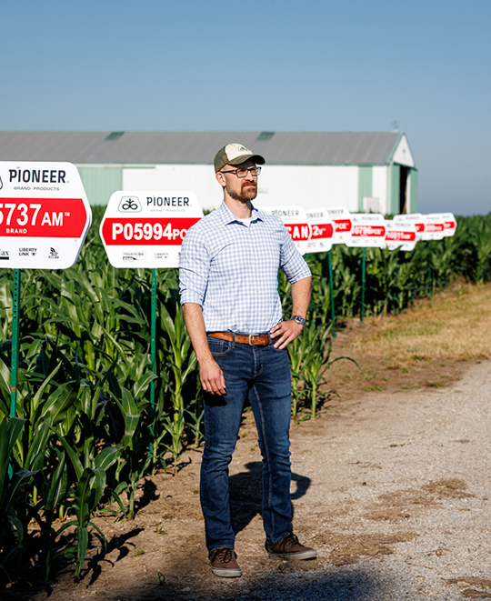 Alumnus Dr. Eric Schultz standing outdoors along a dirt path next to a lush green crop field and a red and white research sign.