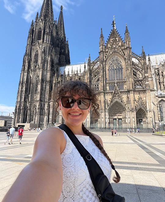 Elizabeth Gruen taking a selfie outdoors with the Cologne Cathedral in the background.
