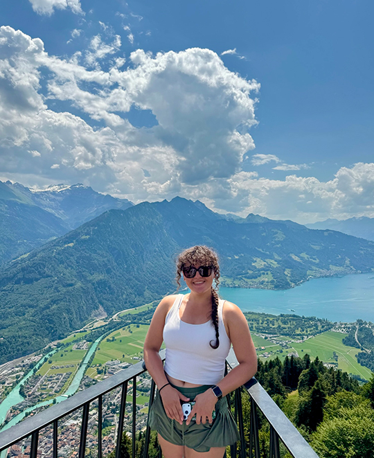 Elizabeth Gruen posing outdoors on a mountain overview during a trip in Switzerland with scenic mountains in the background.